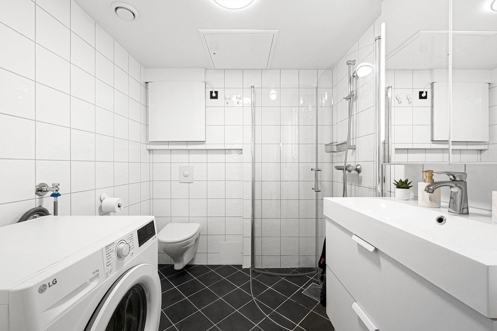 Lyst og moderne bad med praktisk planløsning, flislagte vegger og gulv. Inneholder dusj, servant, vegghengt toalett og vaskemaskin. Galleribilde