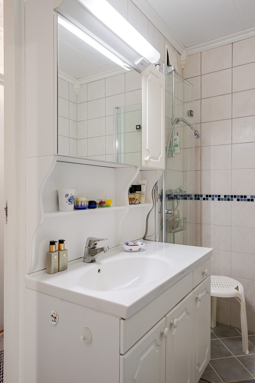 Lys baderomsinnredning med heldekkende servant, speilskap med belysning over. Baderommet er også utstyrt med dusjhjørne med innfellbare glassdører. Galleribilde