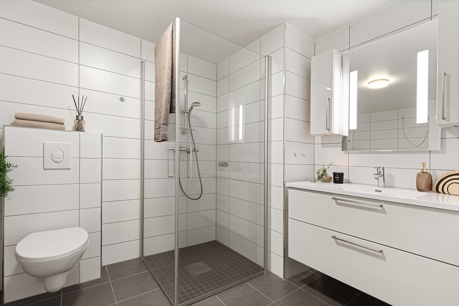 Delikat bad/vaskerom med flislagte gulv og vegger, utstyrt med servantskap med nedfelt servant, veggskap og speil med innebygd belysning. Galleribilde