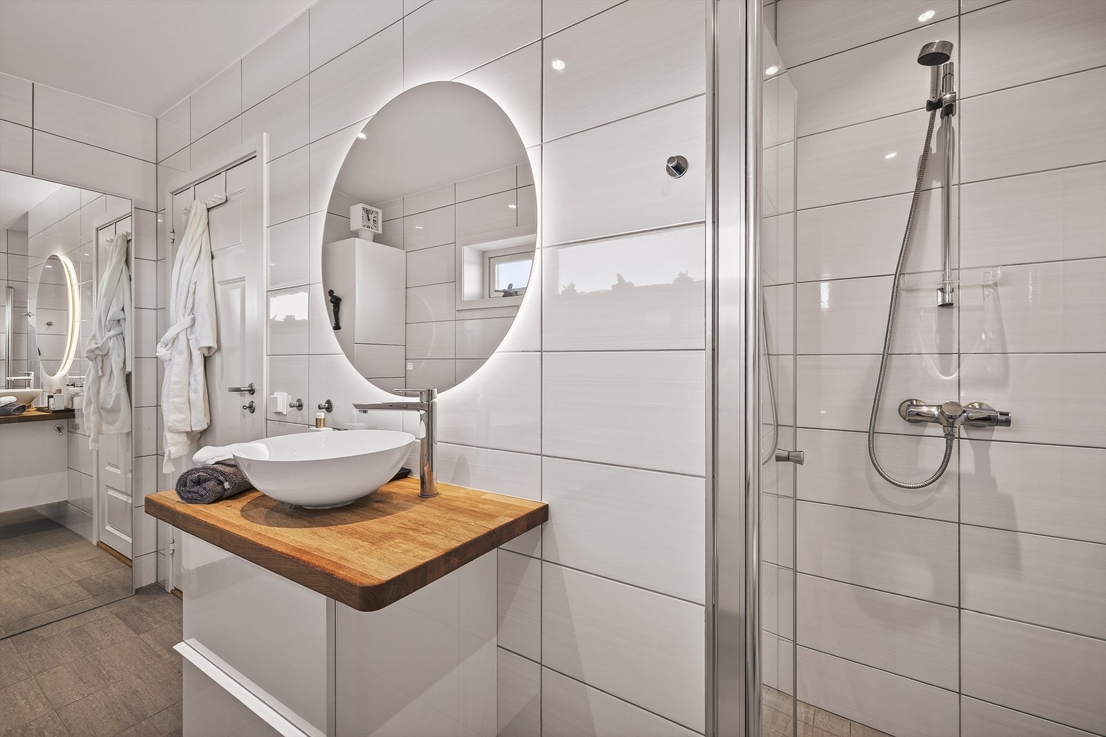 Badet inneholder foldedører i dusj, vegghengt toalett, og baderomsinnredning med servant og belyst speil. Galleribilde