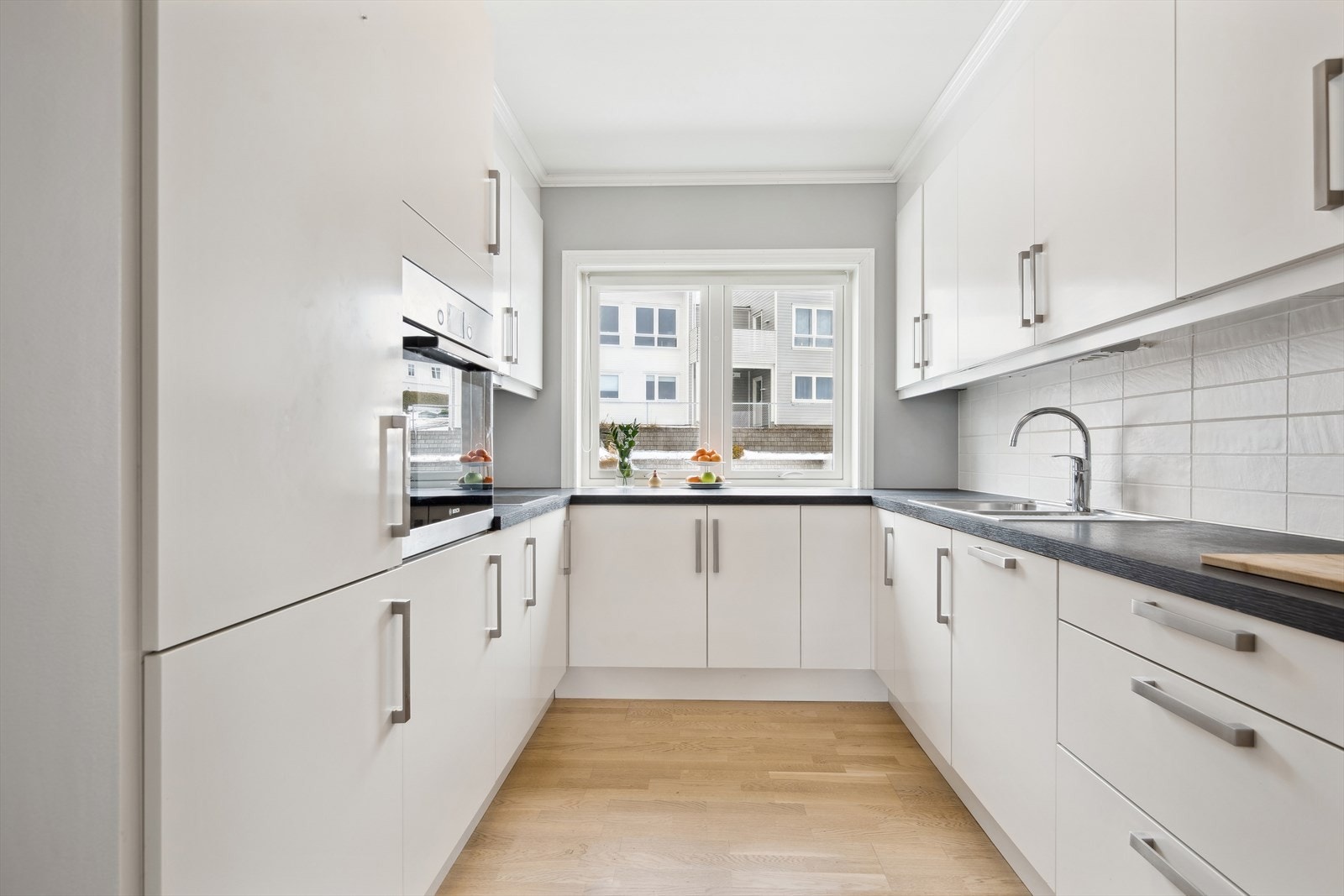 Lys kjøkkeninnredning i hesteskoformasjon med slette fronter, folierte skrog og laminerte benkeplater. Innredningen består av over-/underskap og skuffer. Lyse fliser på veggflatene mellom kjøkkenbenker og overskap. Galleribilde