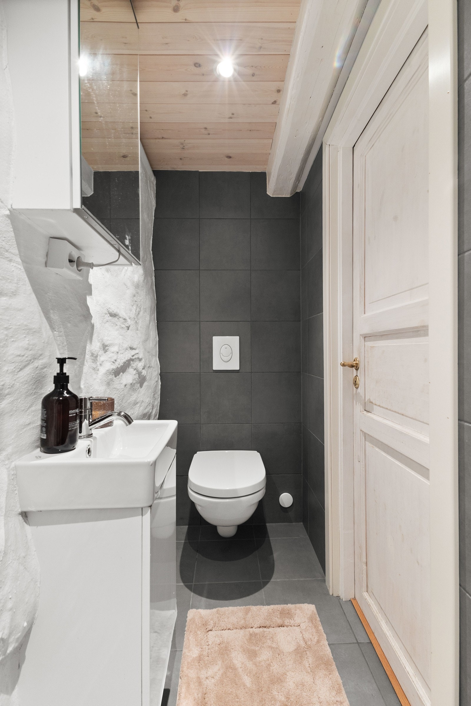 Badet er innredet med regndusj direkte på flislagt gulv, vegghengt toalett, servant og speil, samt spotter i himling. Galleribilde