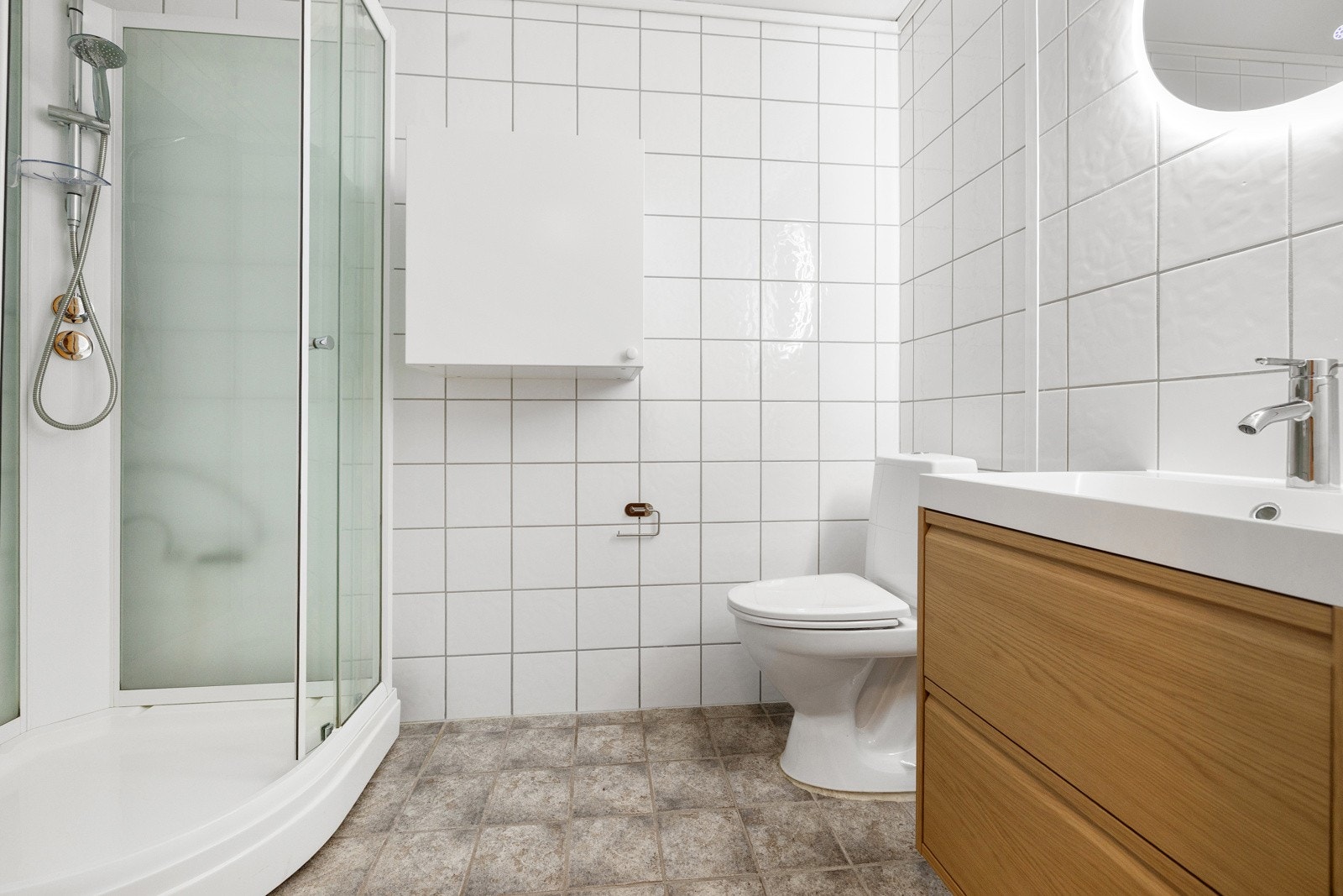 Eldre flislagt bad med med dusjkabinett, toalett og vegghengtskap. Galleribilde