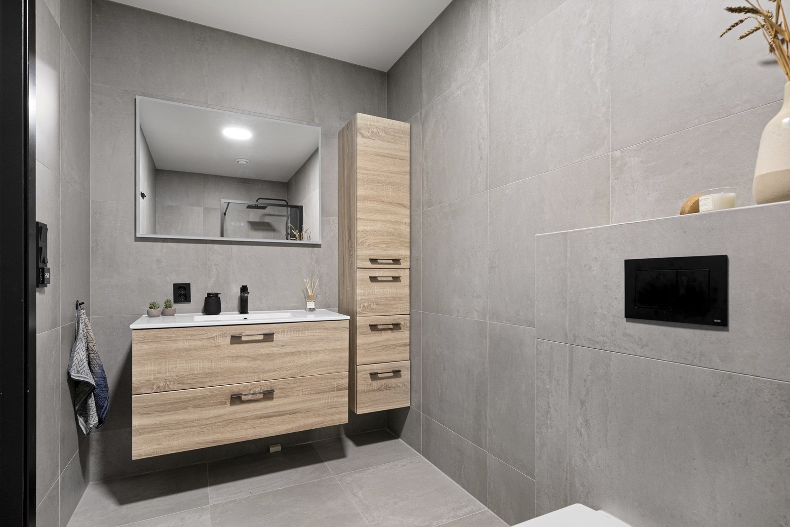 Badet har en moderne finish med store, grå vegg- og gulvfliser. Den øvrige innredningen består av en servant med skuffer, et matchende høyskap og et speil med integrert belysning. Galleribilde