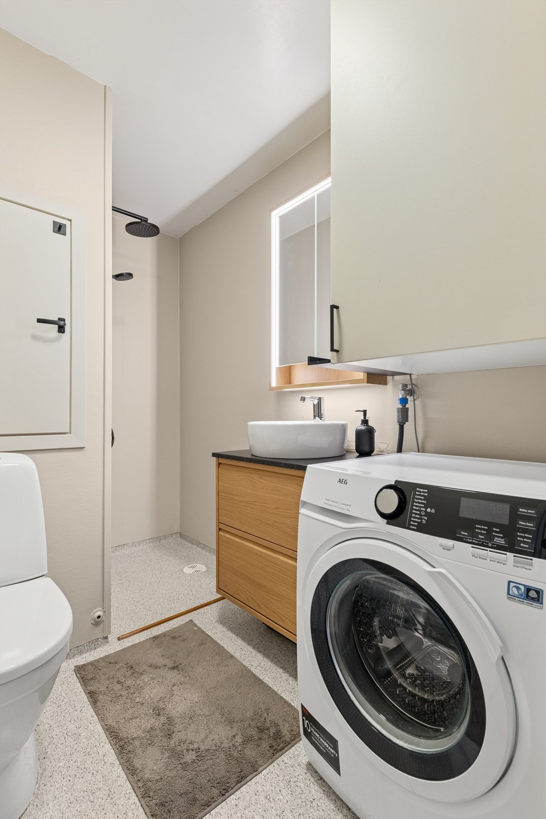 Baderommet er delikat innredet med vinylbelegg på gulv og malt våtromstapet på vegg. I gulvet har man også varmekabler for ekstra komfort. Badet er ellers utstyrt med dusjnisje, toalett, baderomsinnredning med servant og opplegg for vaskemaskin. Galleribilde