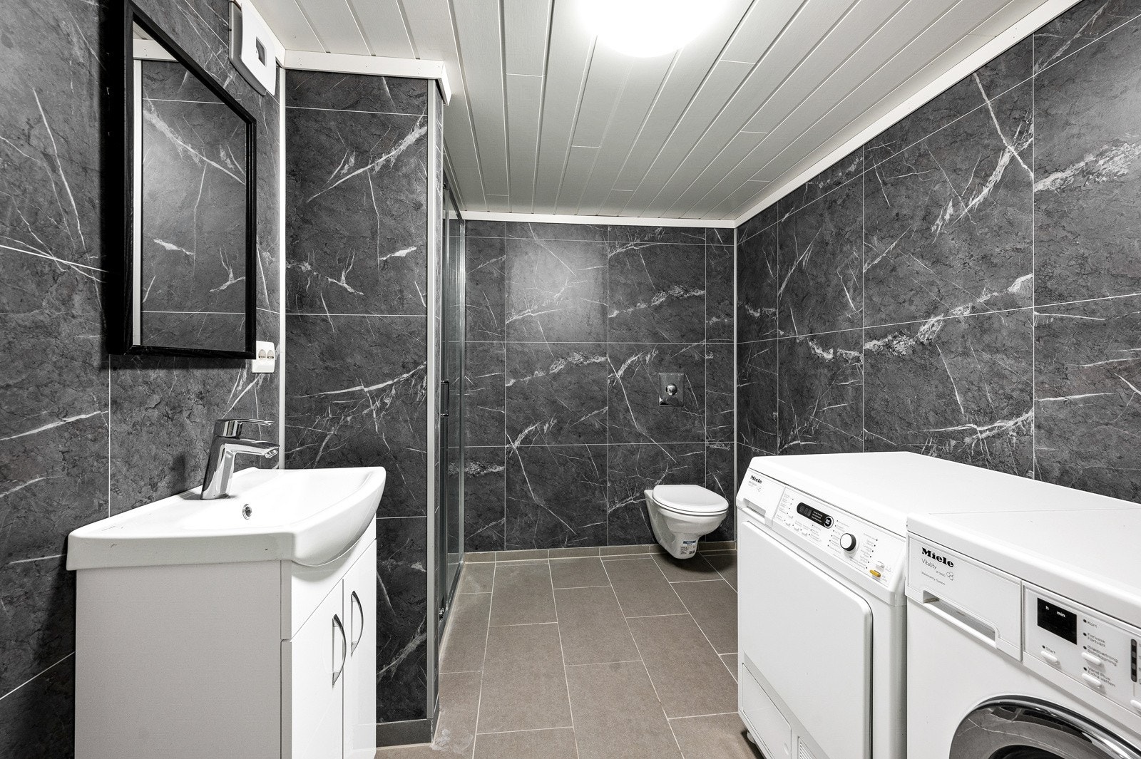 Bad/vaskerom med vegghengt toalett og opplegg for vaskemaskin. Galleribilde