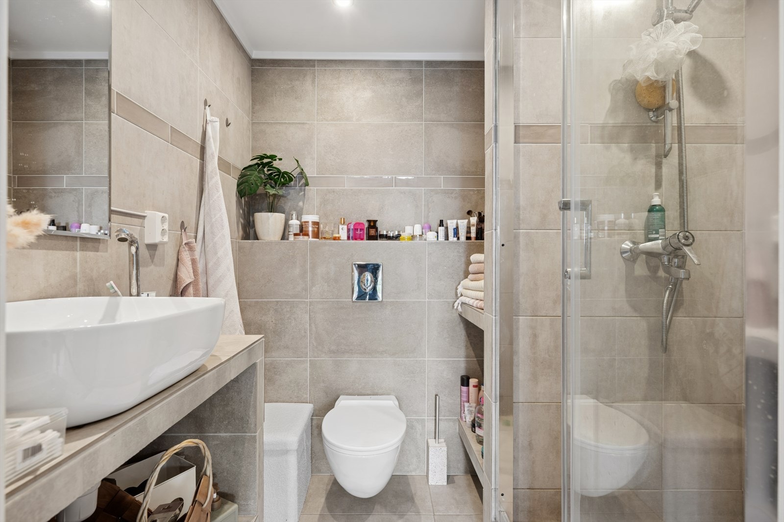 Flislagt bad med speil, servant, veggmontert toalett og dusjhjørne. Galleribilde