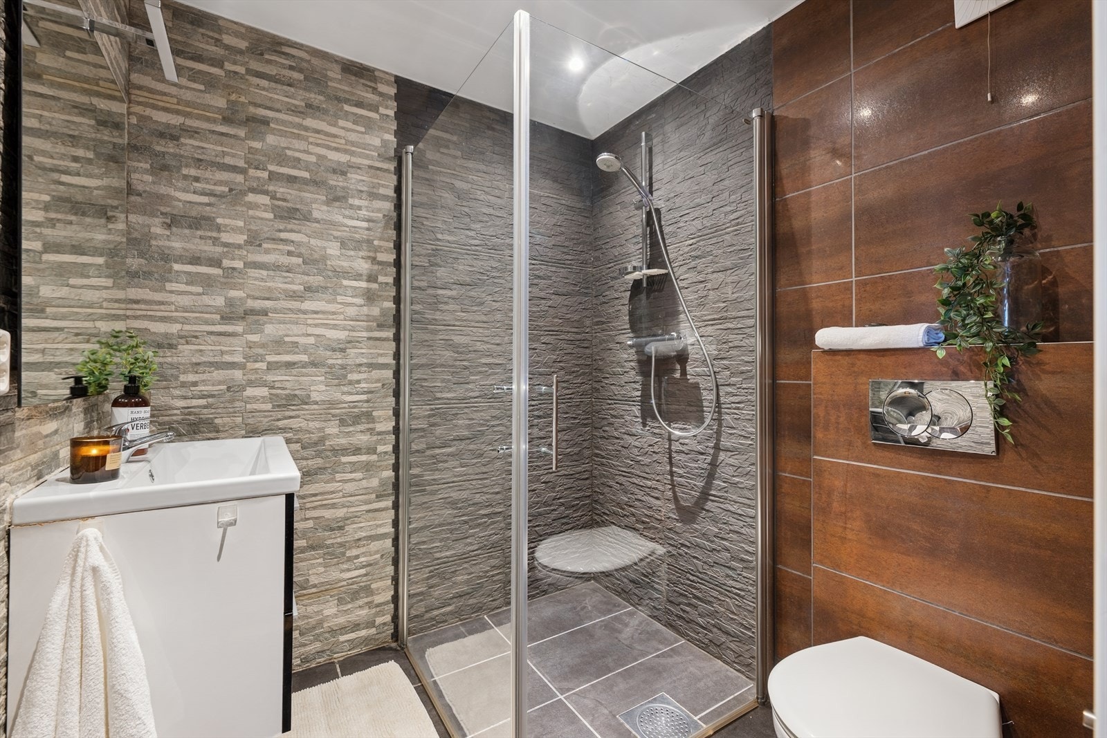 Flislagt bad med speil, servant med seksjon, veggmontert toalett og dusjhjørne. Galleribilde