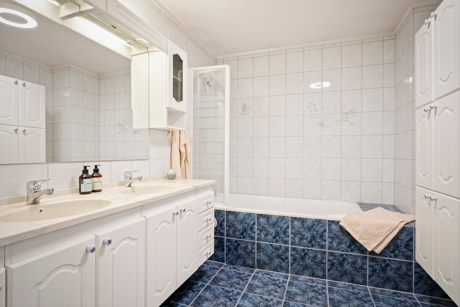 Badet ligger i boligens underetasje og er innredet med fliser på gulv og vegger. Rommet er innredet med baderomsmøbel med heldekkende dobbel servant, profilerte og lyse fronter, samt overhengende speil med belysning. Galleribilde