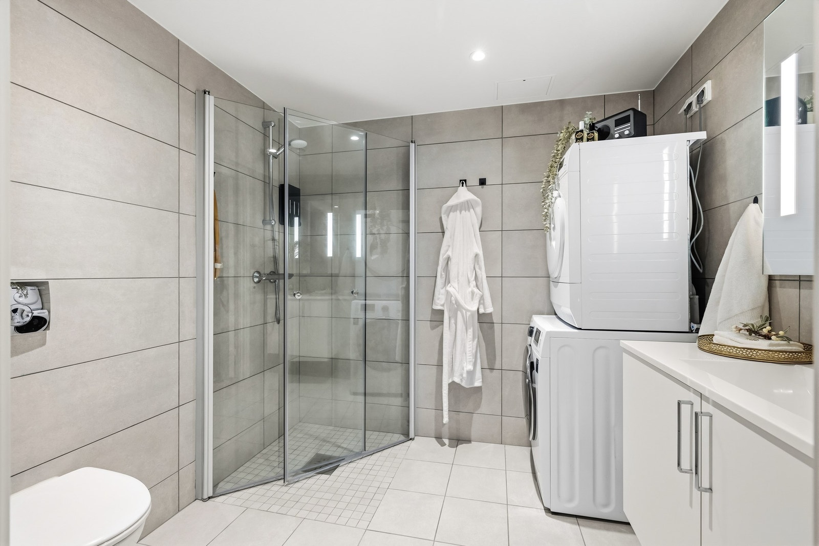 Badet er innredet med veggmontert servantskap med nedfelt servant, dusjhjørne med innfellbare glassvegger, vegghengt toalett samt plass og opplegg for både vaskemaskin og tørketrommel. Galleribilde