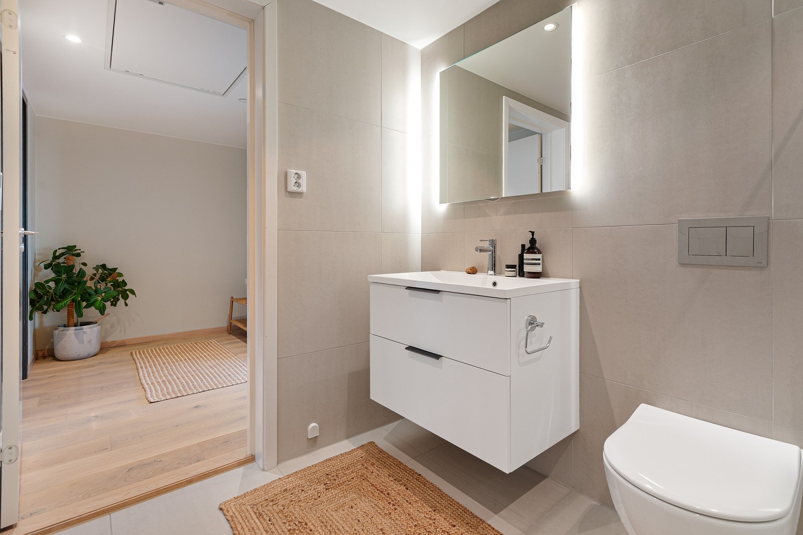 Romslig baderomsinnredning med skuffer og heldekkende servant, samt speil med bakgrunnsbelysning. Badet ligger praktisk tilknyttet entréen med enkel tilgang fra alle rom. Galleribilde