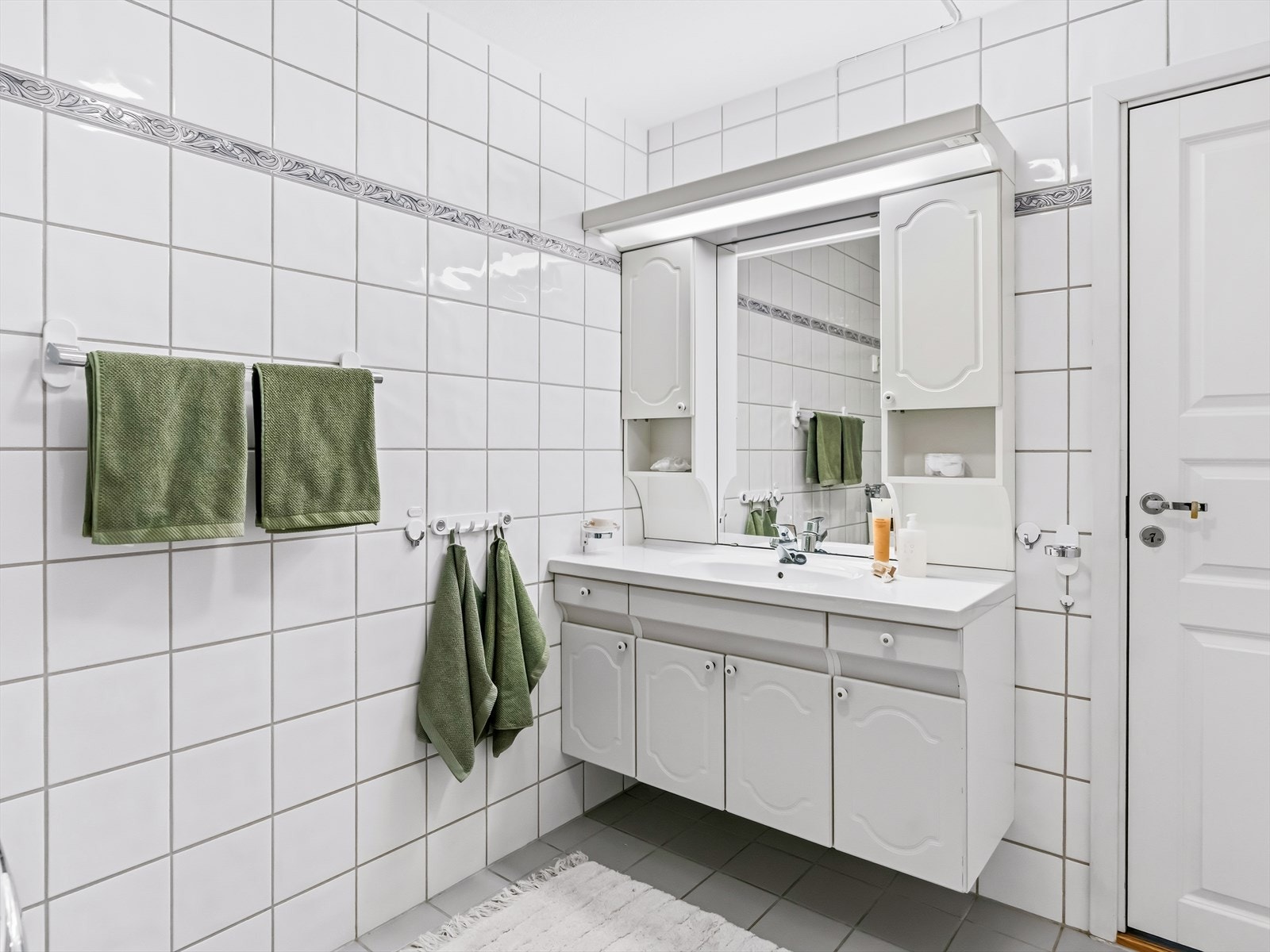 Videre er det servantskap med profilerte fronter og heldekkende servant, samt speil og skap med lysarmatur på veggen over servanten. Det er avtrekk i veggen. Galleribilde