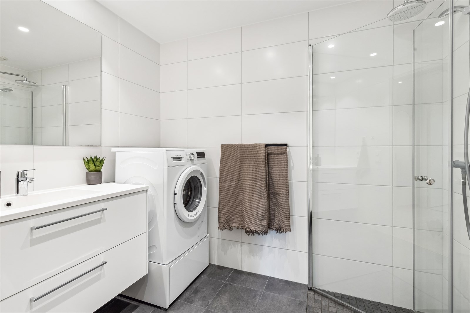 En fin og tidløs baderomsinnredning med hvite, glatte skuffefronter og heldekkende servant. Videre er badet utstyrt med vegghengt toalett, dusj og opplegg for vaskemaskin. Galleribilde