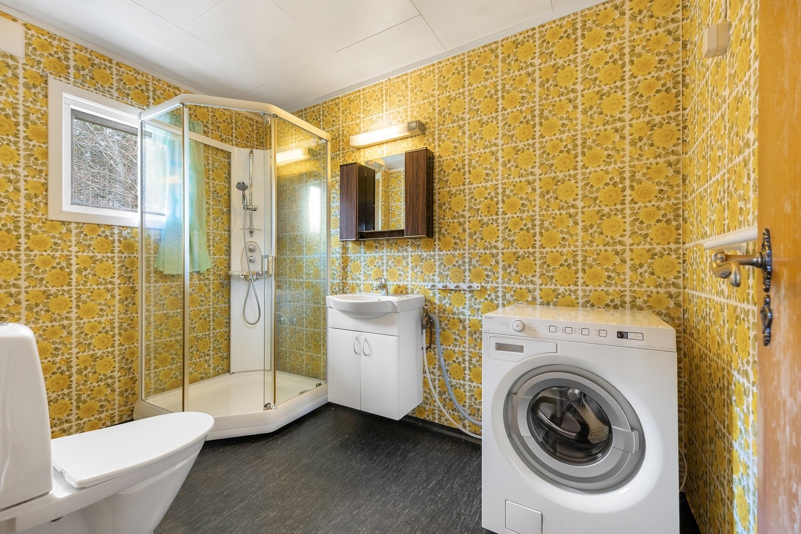 Badet har heldekkende servant over hvitt skap, speilhylle med overlys, dusjkabinett, gulvmontert toalett og opplegg for vaskemaskin. Galleribilde