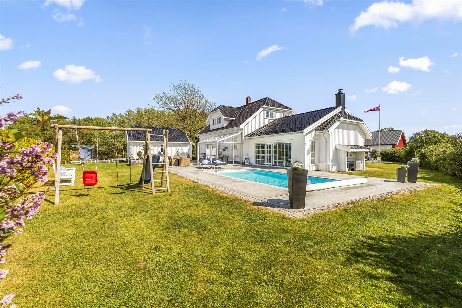 Eiendommen er pent opparbeidet med plen, bassengområde med skiferstein rundt og beplantning med blomster, busker og trær. Galleribilde