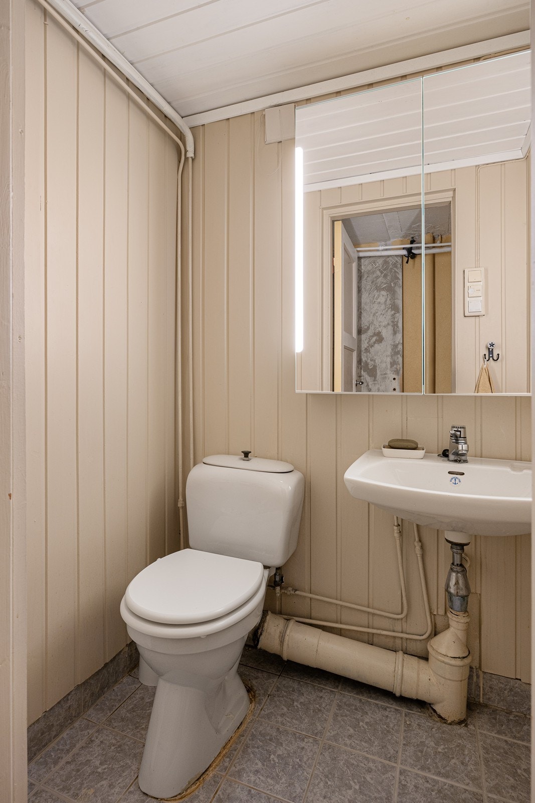 Wc-rommet har panel på veggene og grå fliser på gulvet med lun varme. I tillegg til toalettet består innredningen av en servant og et dobbelt speilskap med integrert belysning. Galleribilde