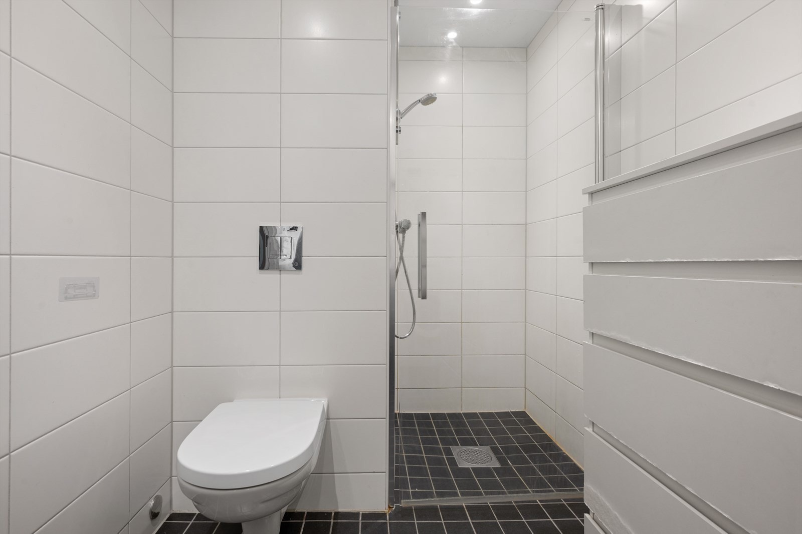 Flislagt bad med vegghengt toalett, dusjløsning og servant på skuffeseksjon. Galleribilde