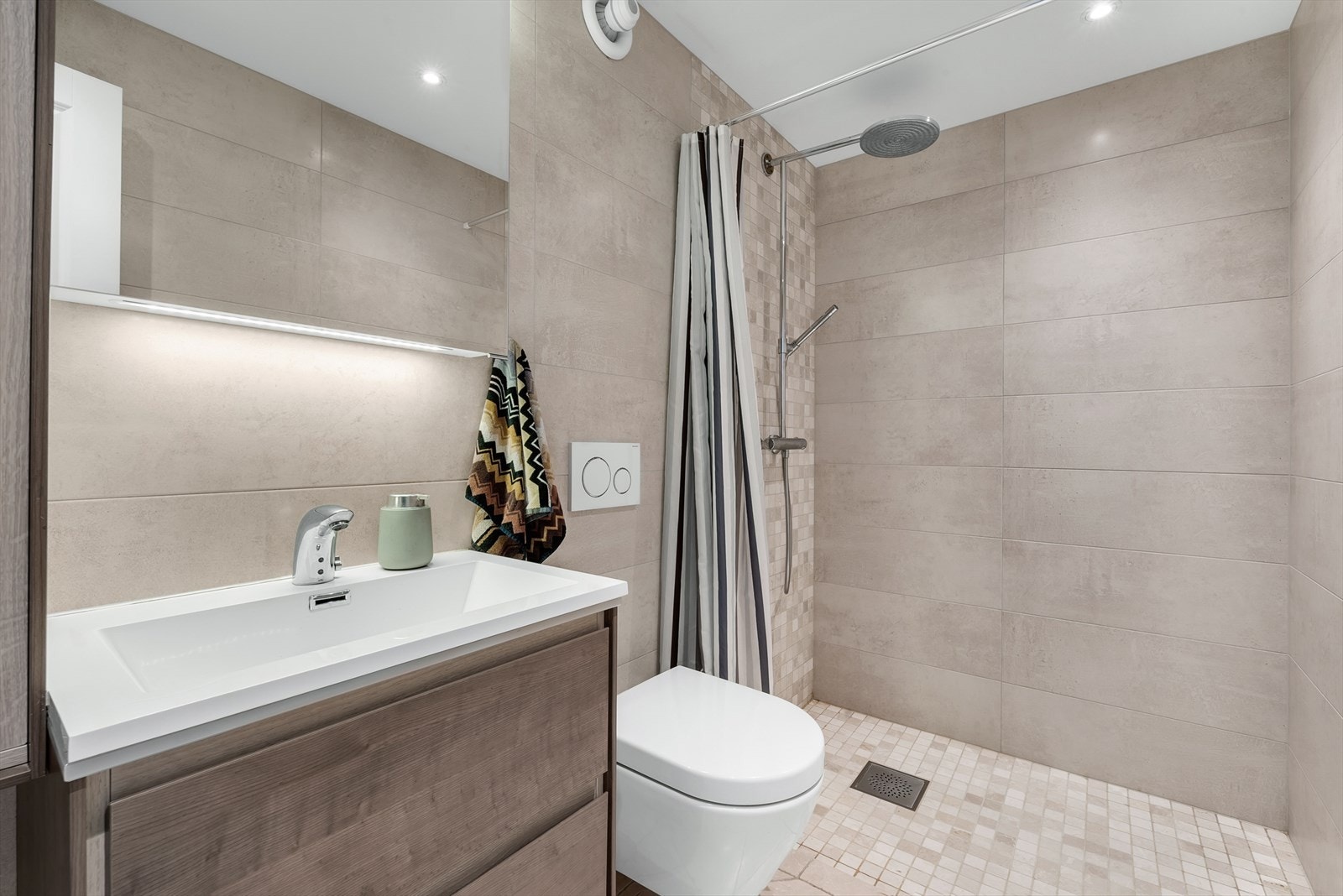 Lekkert bad fra 2017 med flislagte overflater, elektriske varmekabler og spotter i taket. Badet er innredet med nedfelt servant, speil, veggmontert toalett og dusjhjørne. Galleribilde