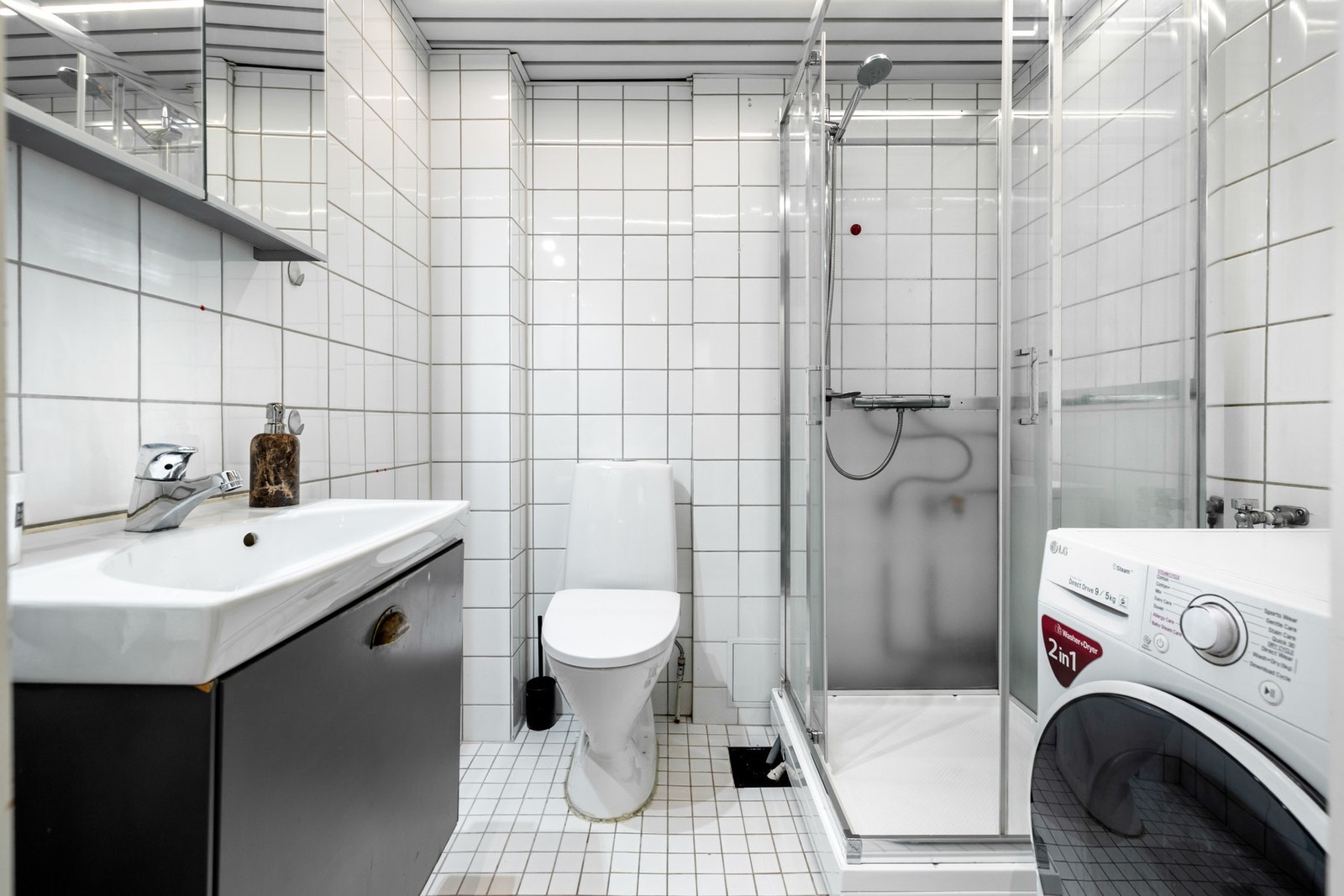 Badet er flislagt med lyse fliser på både vegger og gulv. Rommet er innredet med servant med skuffe og speil med skap, som gir praktisk oppbevaring. Galleribilde