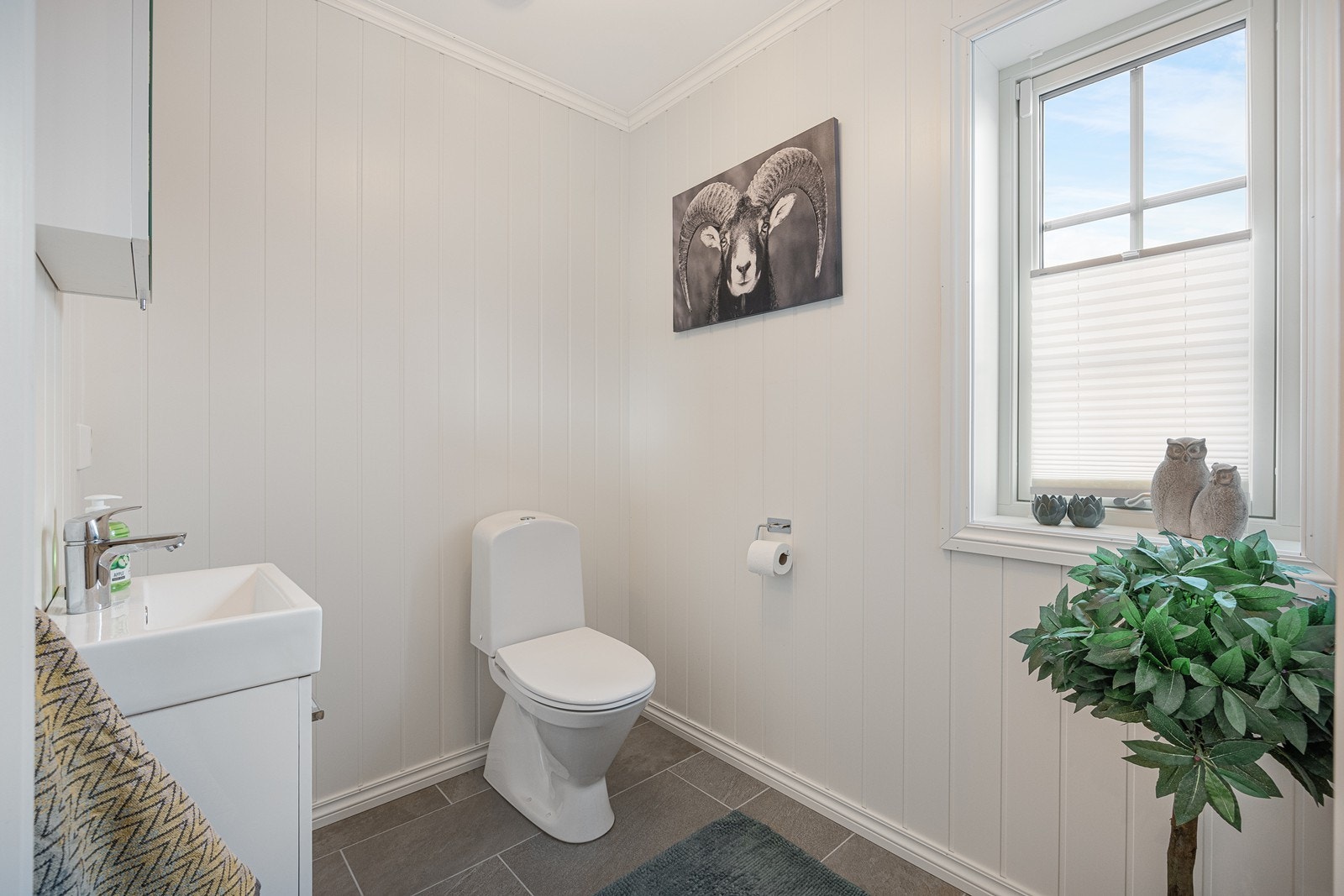 Gjestetoalettet ved entreen har fliser på gulv og malte plater på vegger og tak. Rommet er innredet med gulvstående toalett og enkelt servantskap. Galleribilde