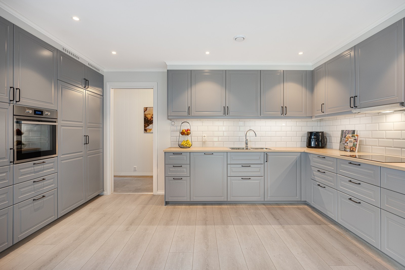 Kjøkkeninnredningen er fordelt på to takoppbygde seksjoner i klassisk stil, med profilerte, grå fronter som passer til øvrig fargevalg. Benkeplaten er i laminat og matcher gulvet og har hvite fliser i bakkant, samt benkebelysning. Galleribilde