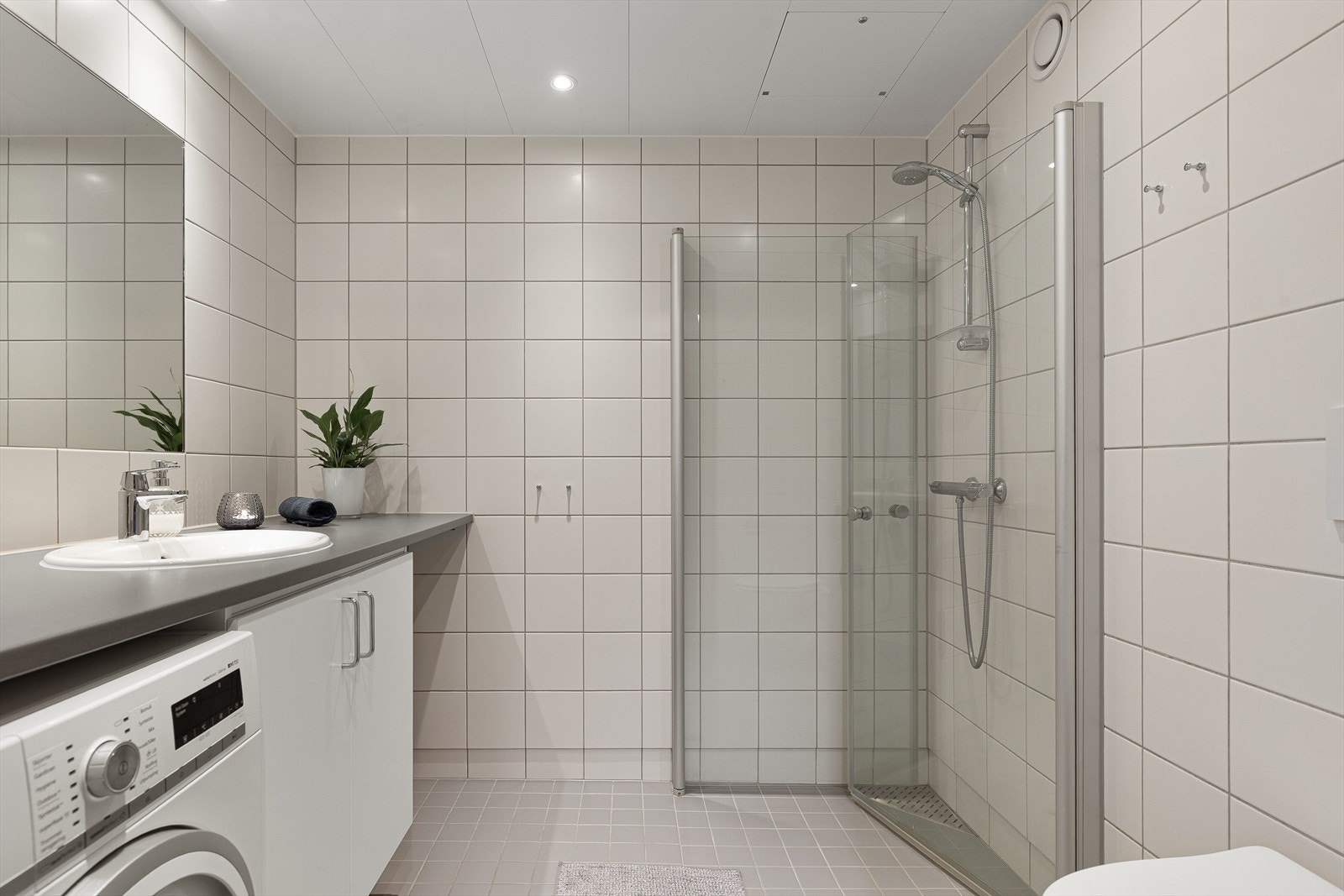 Bad/vaskerommet er innredet med laminat benkeplate, servantskap med nedfelt servant, innmurt speil, vegghengt toalett med innebygd sisterne samt dusjhjørne med innfelte dusjvegger i herdet glass. Galleribilde