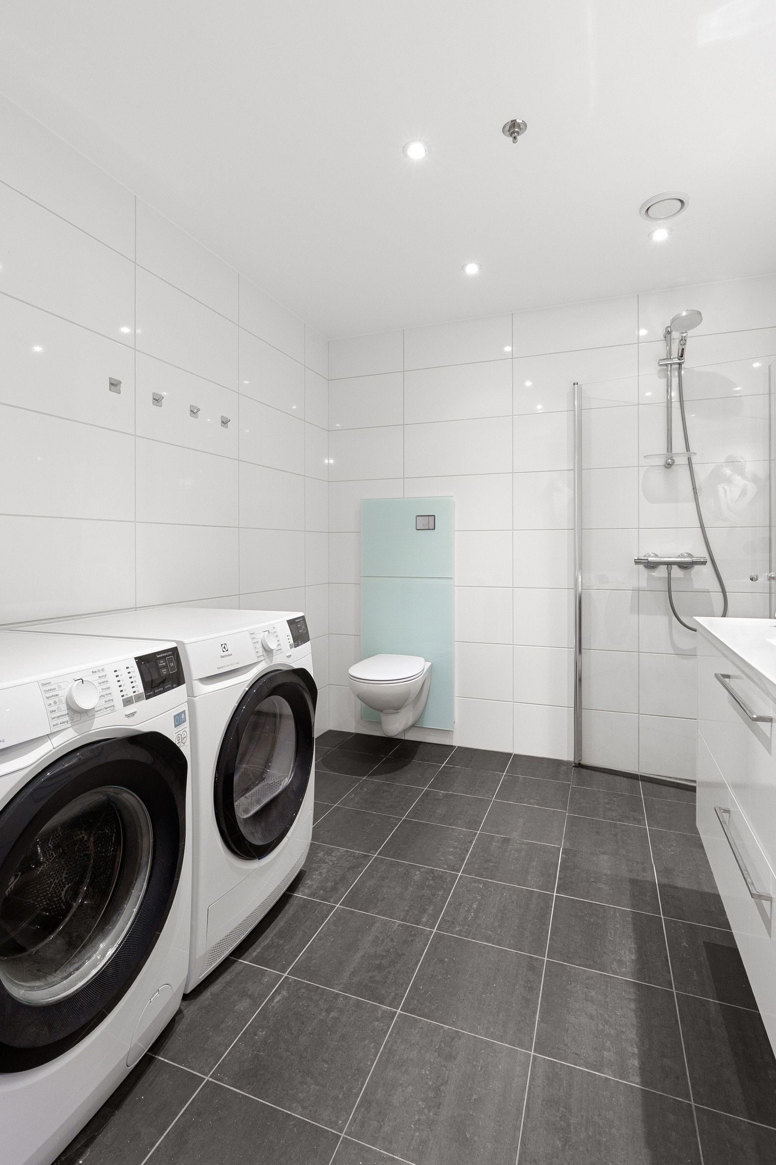 Bad med dusjhjørne med glassdører, vegghengt toalett og servantinnredning med skuffer. Opplegg for vaskemaskin og tørketrommel. Galleribilde