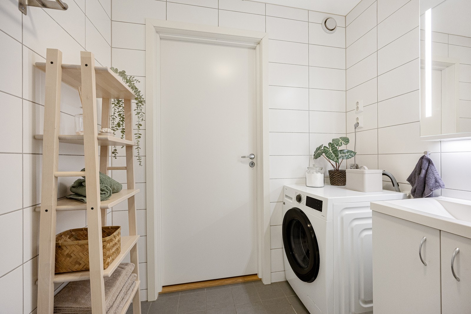 Badet har en fin størrelse, innredet med hvite veggfliser og lysegrå gulvfliser i ulike størrelser. Innredningen består av et hvitt servantskap og et tilhørende speilskap med integrert belysning. Galleribilde