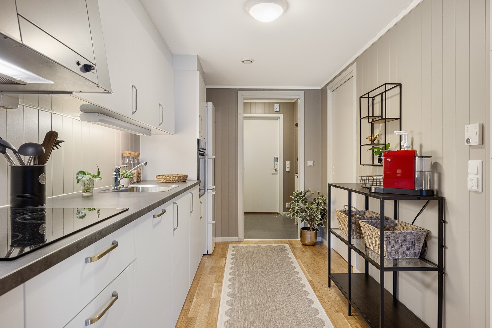 Kjøkkeninnredningen har hvite, glatte fronter, nedfelt oppvaskkum og grå benkeplate av laminat. Belysning er montert over benken, og over kokesonen har kjøkkenet en hetteventilator med avtrekk. Galleribilde