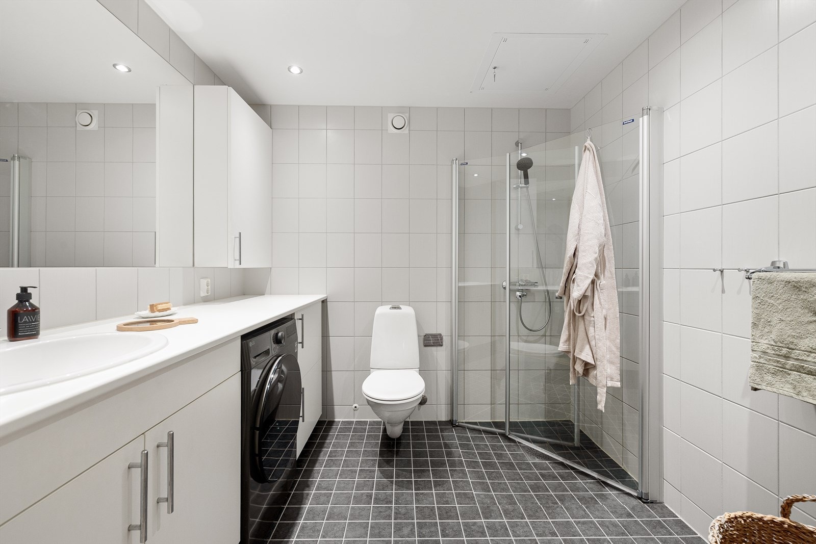 Stort hovedbad med vegghengt toalett, opplegg for vaskemaskin, innfellbare dusjdører, speil, overskap og elektriske varmekabler i gulv. Galleribilde