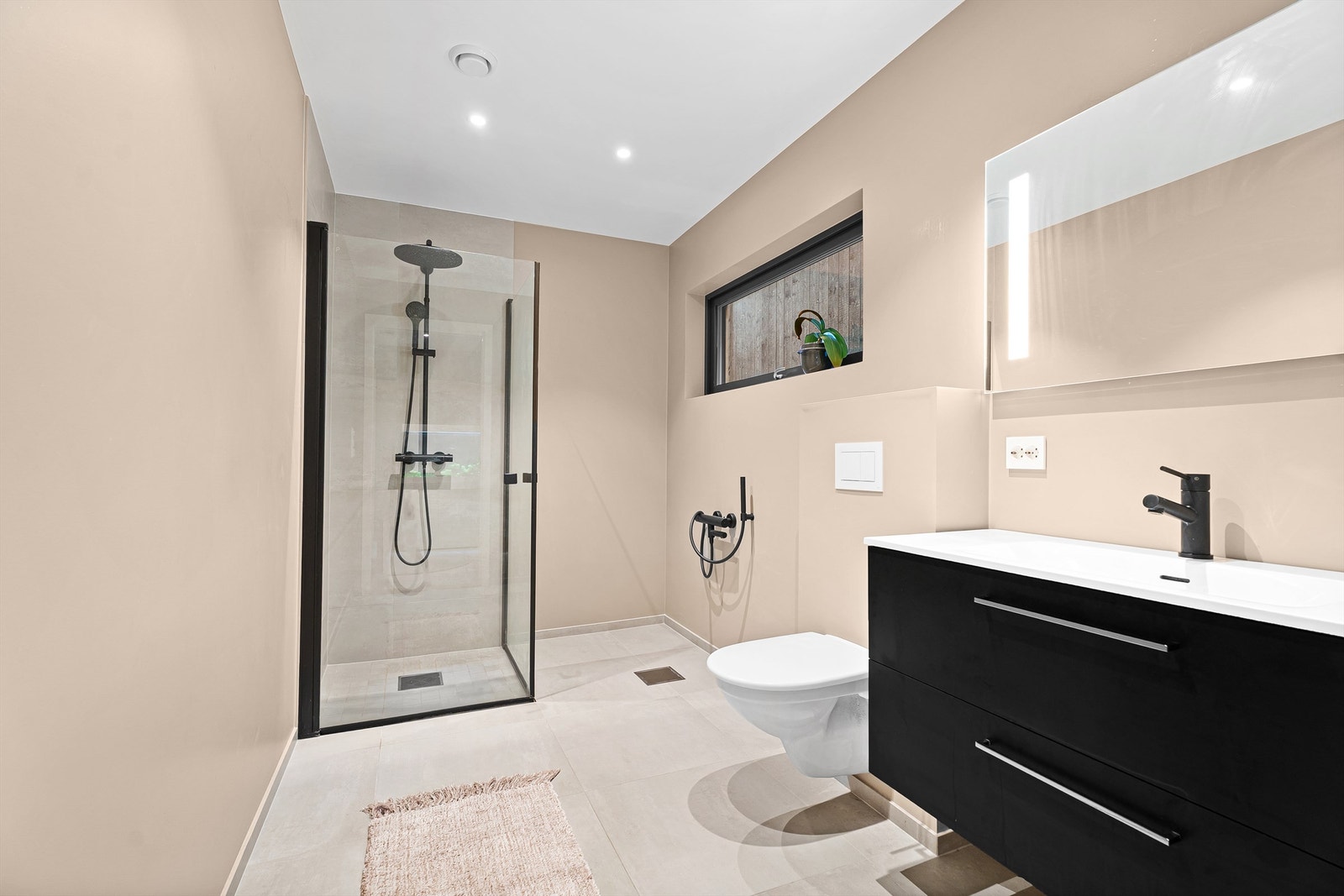 Badet er romslig og har fliser på gulv og i dusjsonen. Videre er det vegghengt toalett samt servant med oppbevaring og speil. Galleribilde