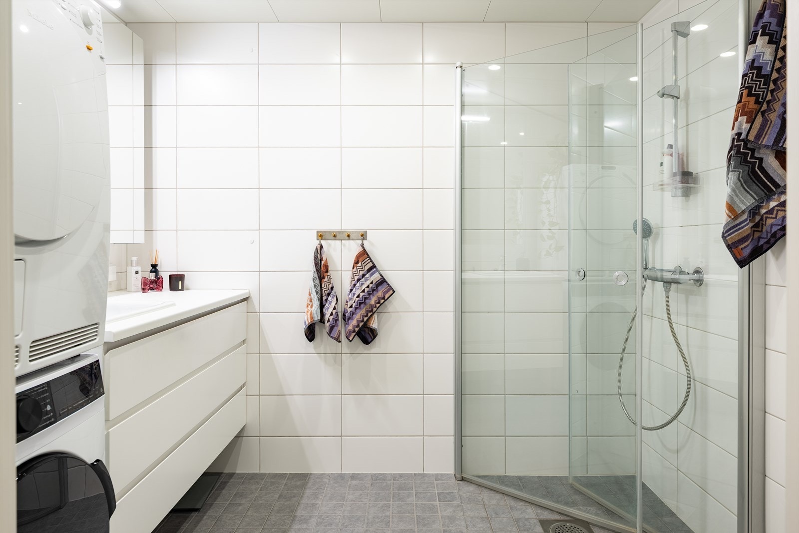 Delikat baderom med fliser på gulv/vegg, servant med underskuffer, overskap med speil, dusjhjørne med innfellbare dusjdører og vegghengt toalett. Galleribilde