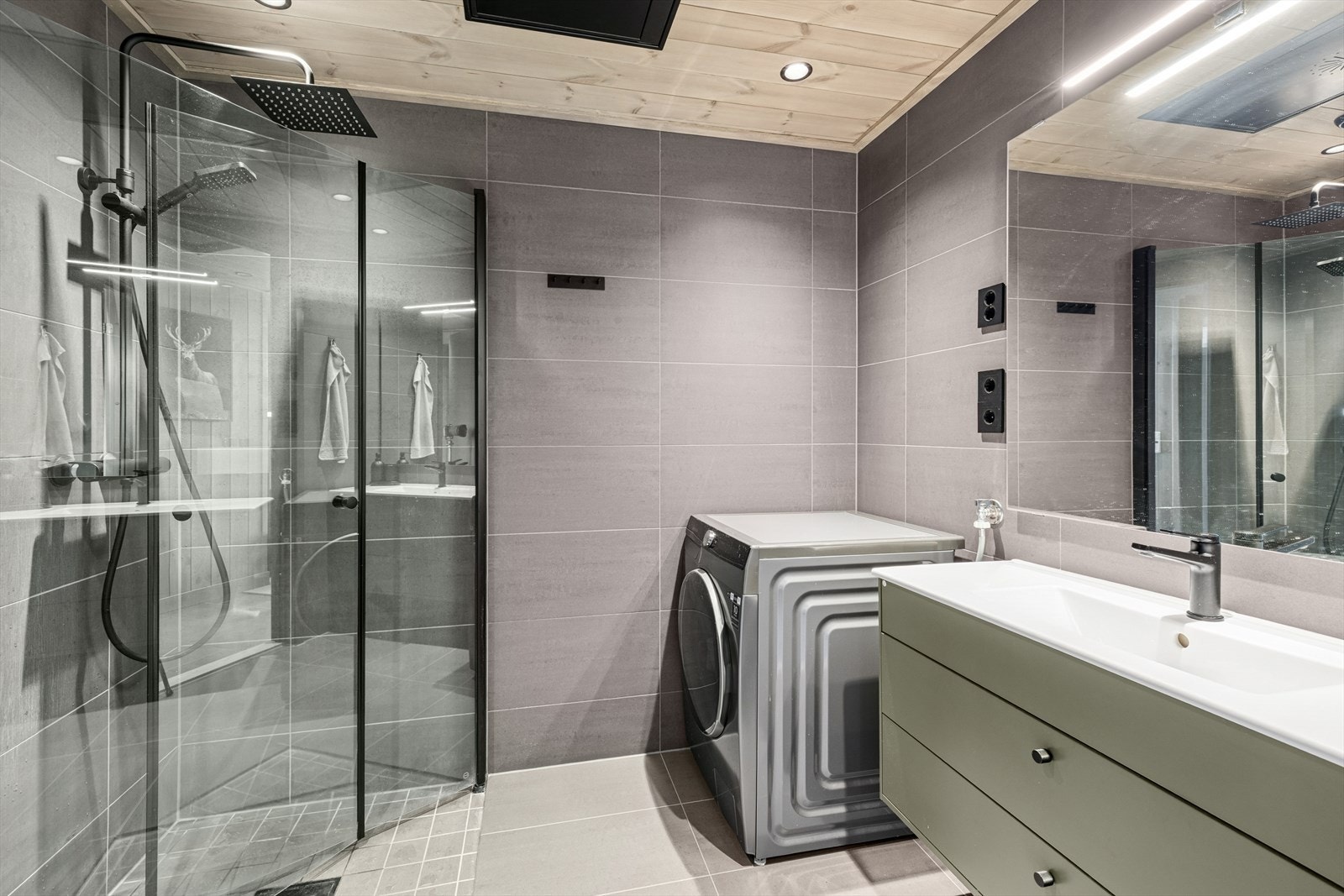 Badet er utstyrt med servant med underskap, speil med tilhørende belysning, dusjvegger, vegghengt wc, og opplegg for vaskemaskin. Galleribilde