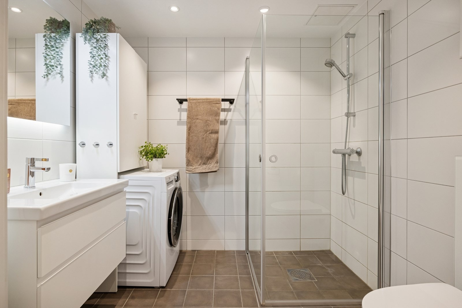 Flislagt baderom utstyrt med dusj, opplegg for vaskemaskin og tørketrommel, vegghengt toalett og servant med underskap og speil. Galleribilde