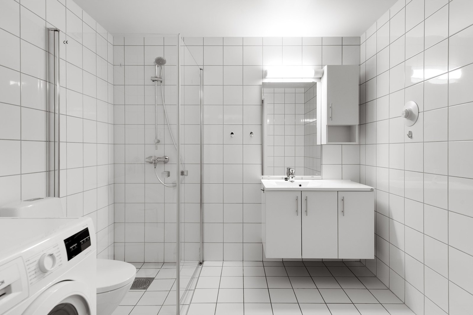 Pent helfliset baderom med tidløse flisvalg. Servant i innredningen, dusjhjørne og opplegg for vaskemaskin. Galleribilde