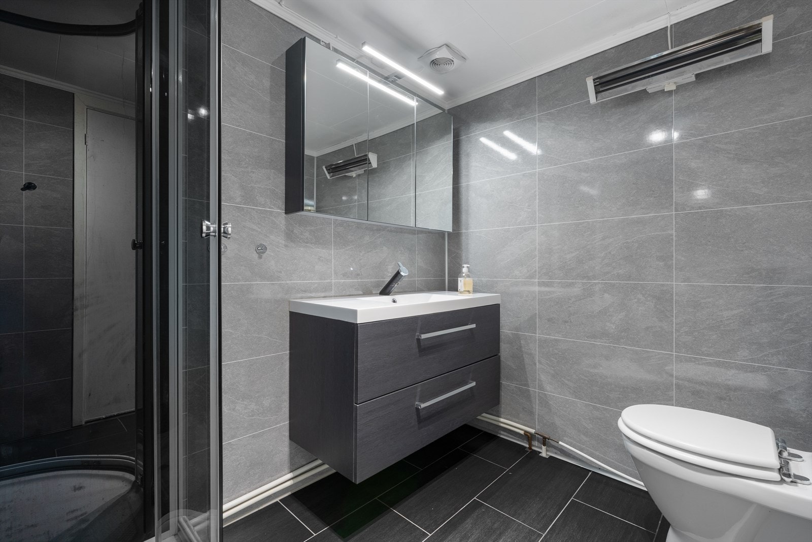 Moderne bad med stilrene grå fliser og elegant servant. Praktisk dusjløsning og god belysning gir et funksjonelt og innbydende rom. Galleribilde