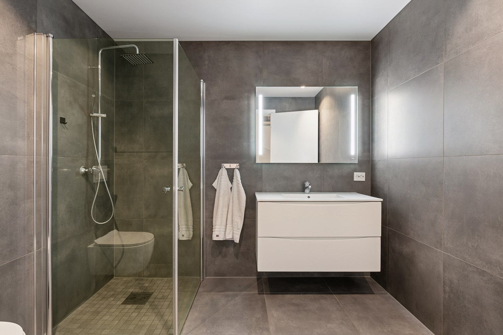 Delikat, flislagt baderom innredet med vegghengt toalett, dusjhjørne og baderomsinnredning med nedfelt servant. Galleribilde