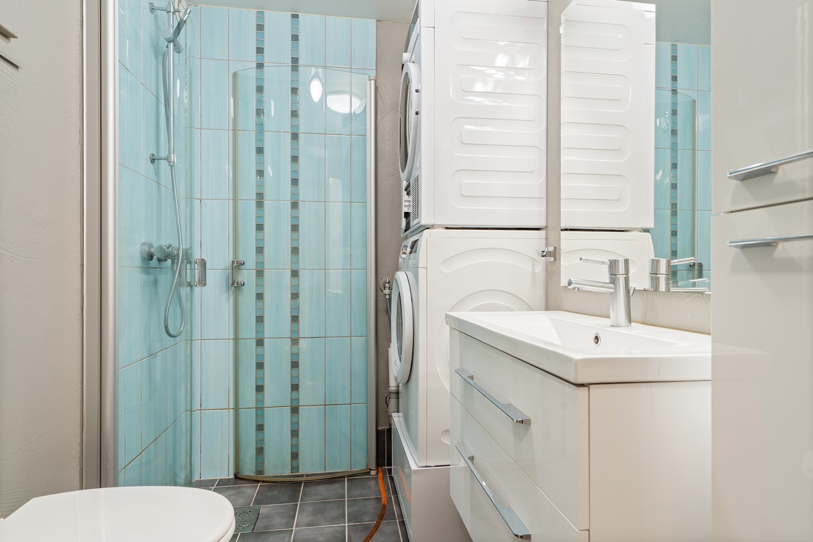 Bad med fliselagt gulv med varmekabler, dusjnisje m/flislagte vegger, vegghengt toalett, vaskemaskin og tørketrommel som medfølger kjøpet, moderne servant med dobbel underskuff og høyt sideskap. Galleribilde