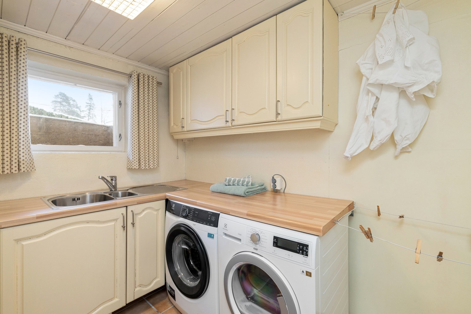 Vaskerom med plass for vaskemaskin og tørketrommel under praktisk brettebenk. Benkeskap med dobbel vask og veggskap til oppbevaring av vaskemidler og lignende Galleribilde