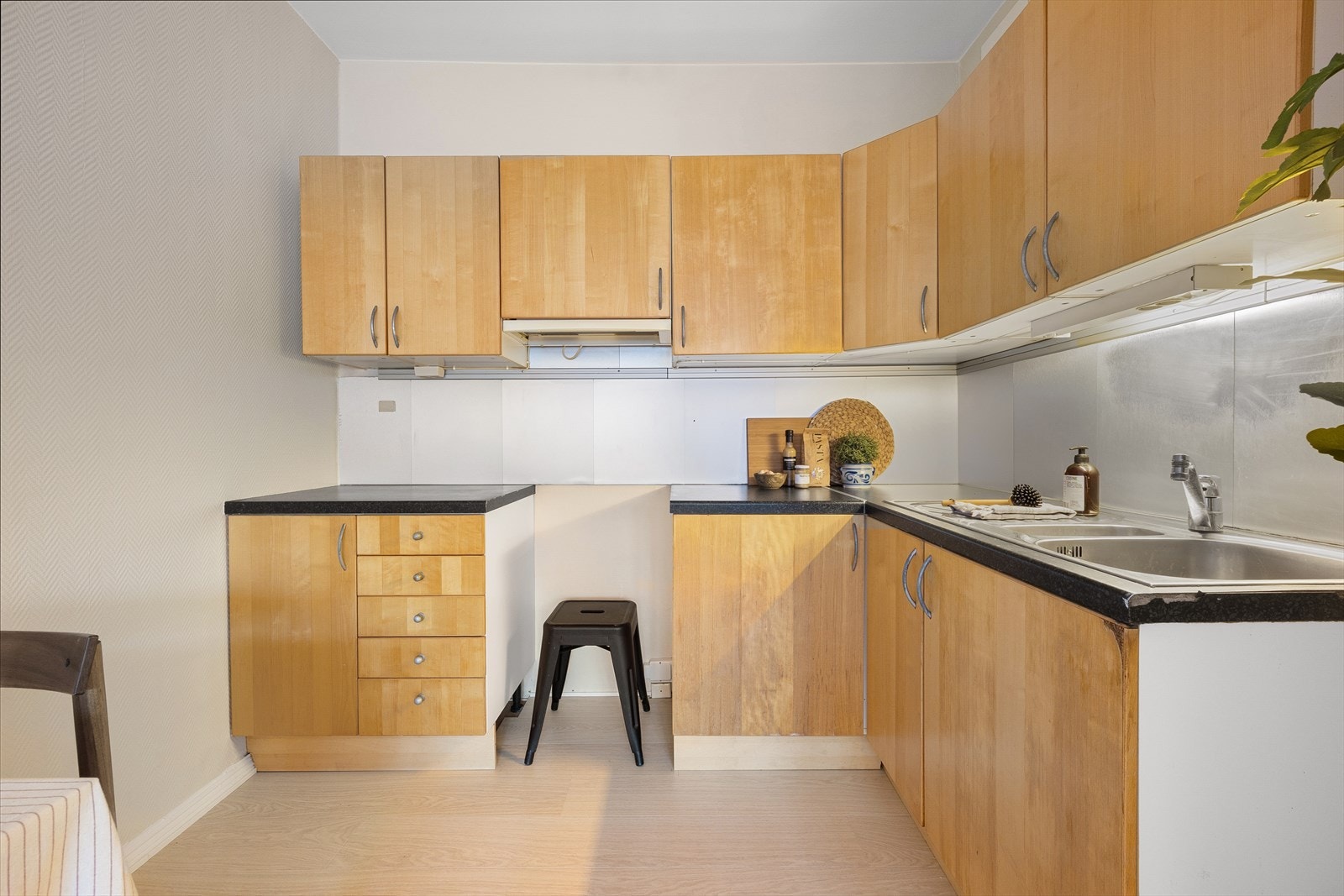 Kjøkkeninnredningen har glatte fronter. Laminat benkeplate med nedfelt kum i rustfritt stål. Stålplate på vegg over benkeskap. Kullfilterventilator. Galleribilde