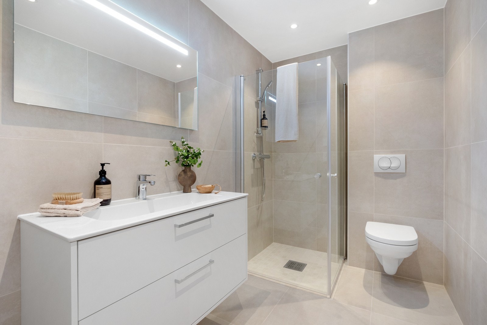 Badet er romslig og moderne, innredet med vegghengt toalett, dusj med foldedører i glass, servant med møblement, speil og spotter i tak. Galleribilde