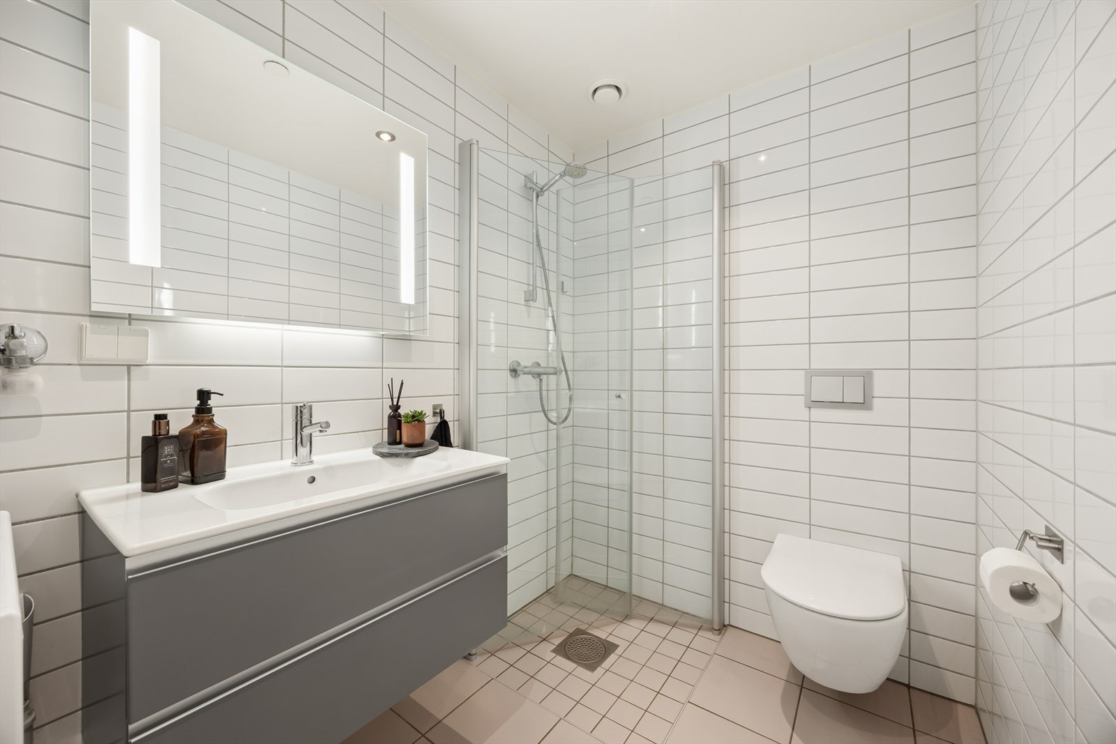 Pent, flislagt baderom utstyrt med innredningen med nedfelt servant, veggmontert toalett og et dusjhjørne med dusjvegger. Galleribilde