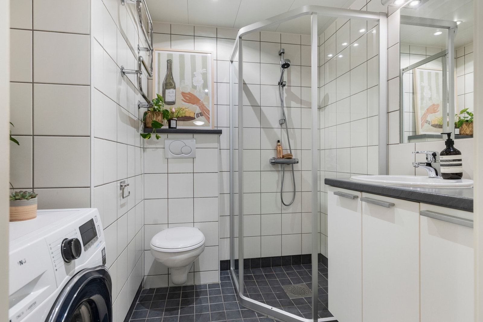 Prefabrikert våtrom fra byggeår med varmekabler, flislagte flater, dusjhjørne, veggmontert servantinnredning og toalett samt opplegg for vaskemaskin og elektrisk håndkletørker Galleribilde