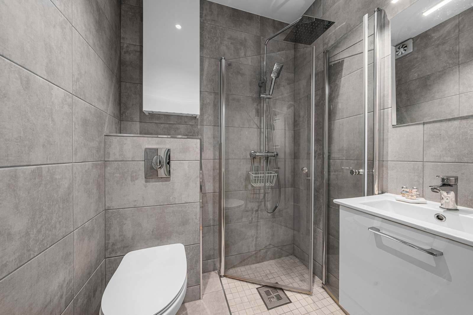 Stilrent bad/WC med flislagte gulv og vegger. Sparklet og malt himling med innfelte downlights. Rommet har nedsenket dusjhjørne med glassdører i herdet glass, vegghengt toalett samt heldekkende servant med underskap og speil med integrert belysning Galleribilde