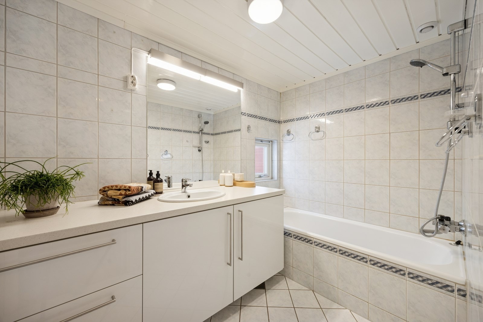 Badet er utført med flislagte overflater i nøytrale farger og har elektriske varmekabler i gulvet - Her er det innebygd badekar m/dusjgarnityr på vegg og baderomsinnredning med mye oppbevaringsplass Galleribilde