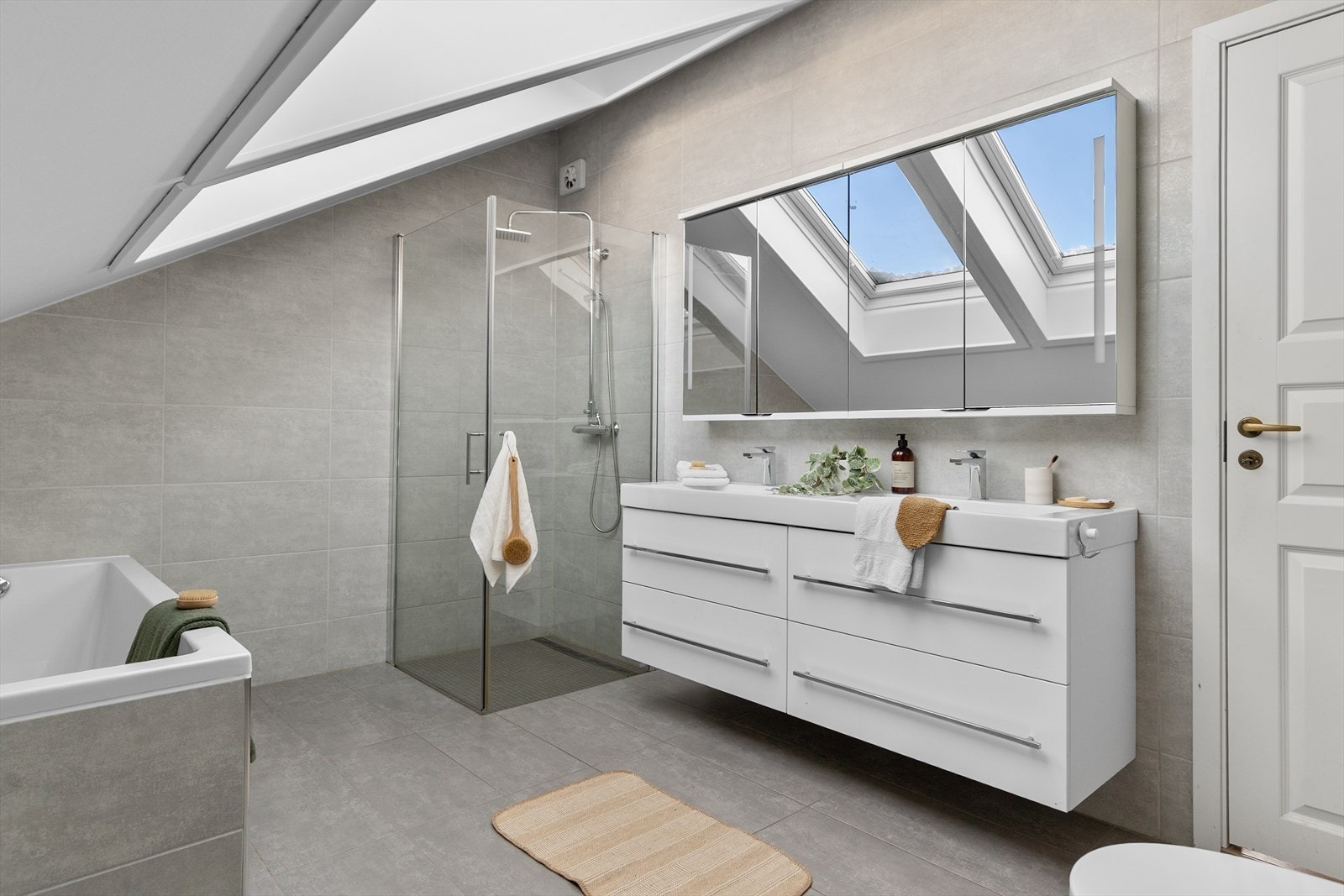 Badet er innredet med dobbel servant med overskap med speilfront og stikkontakt, veggmontert toalett, dusjhjørne med glassdører og et herlig innfliset badekar. Galleribilde