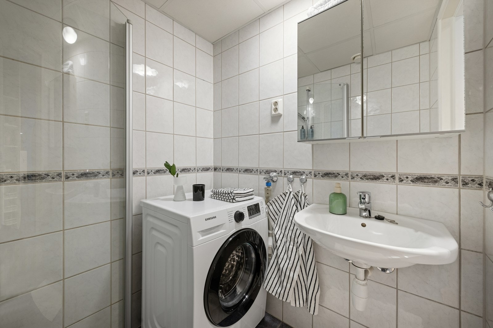 Innredet med dusjvegger med dusjgarnityr på vegg, opplegg for vaskemaskin, vegghengt toalett, servant og overskap. Galleribilde