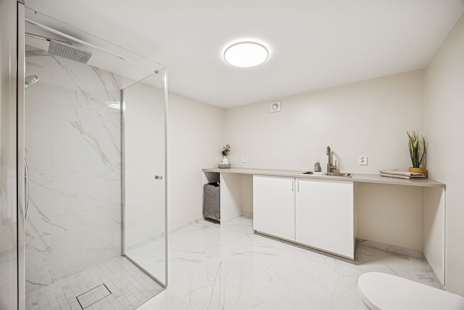 I tilknytning til hoveddelen i kjelleren er det et praktisk bad/vaskerom. Rommet er utstyrt med en romslig dusj med glassvegger og regndusjhode, samt en praktisk benk med integrert vask og hvite skapfronter som gir god oppbevaringsplass. Galleribilde