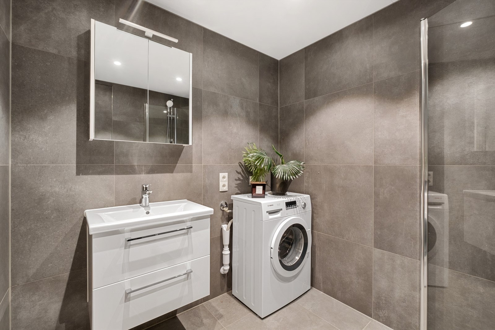 Rommet er innredet med dusjhjørne med innfellbare glassdører, veggmontert toalett og servant med hvit skuffinnredning under samt speil med belysning over Galleribilde