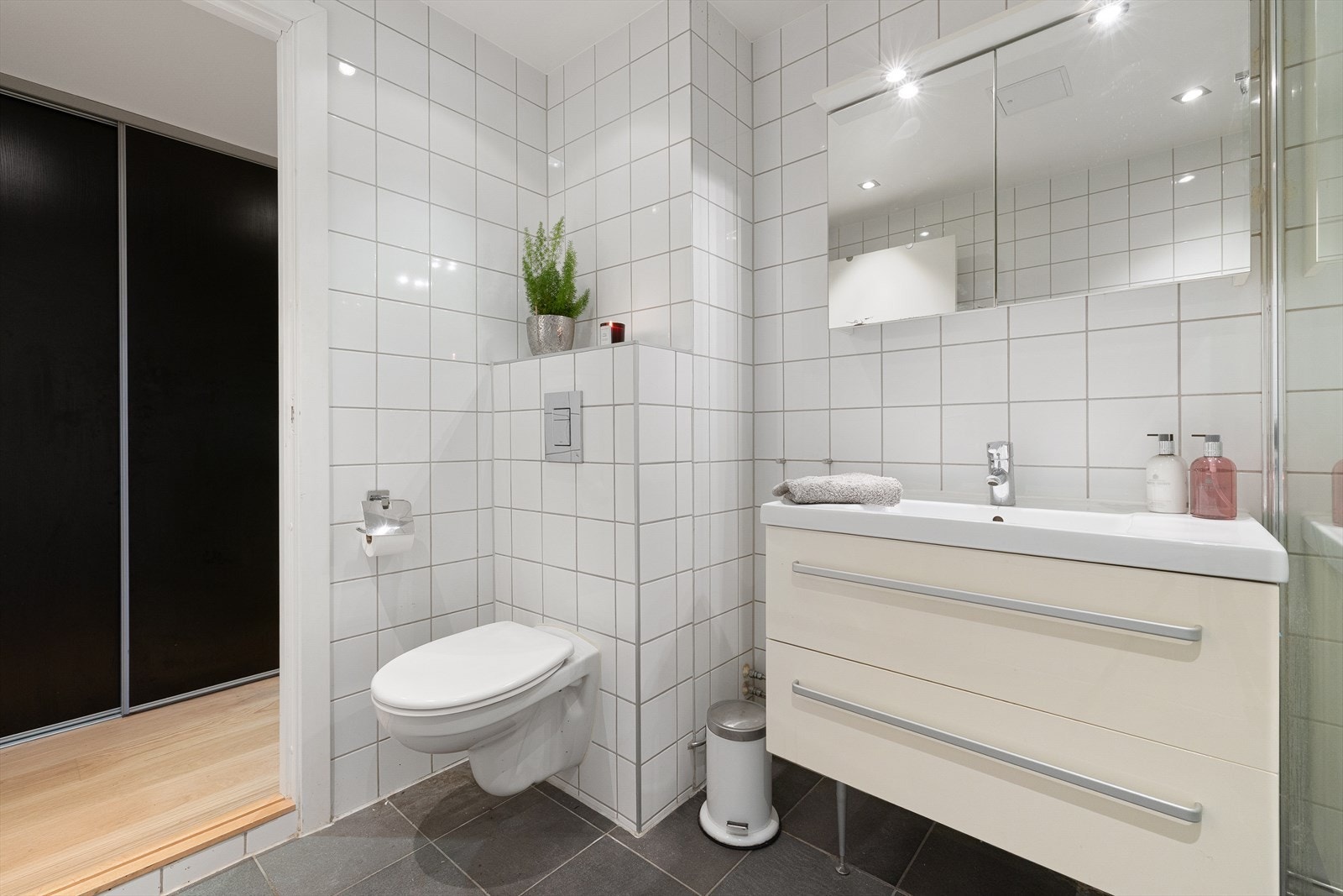 Badet er utstyrt med dusjhjørne med glassvegg, vegghengt toalett og baderomsinnredning med skuffer, servant og vegghengt speilskap. Galleribilde